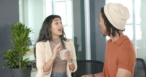 Indian woman and coworker animatedly chatting over coffee in modern collaborative office