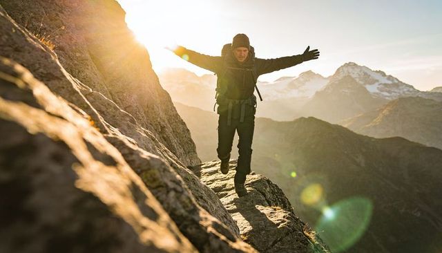 Hiker balancing on narrow alpine ledge at golden hour with backpack and lens flare