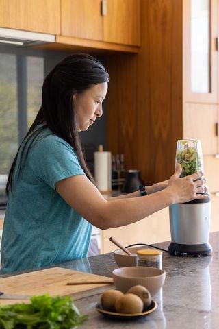 Asian woman blending green smoothie at modern kitchen counter with kiwi and leafy greens