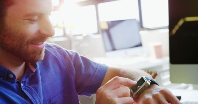 A young man wearing a blue shirt smiles while interacting with his smartwatch in a contemporary office setting. This image is suitable for depicting modern business environments, the integration of technology in daily work life, and promoting wearable tech or productivity apps. Ideal for websites, blogs, advertisements related to business technology, innovative work solutions, and tech advancements.