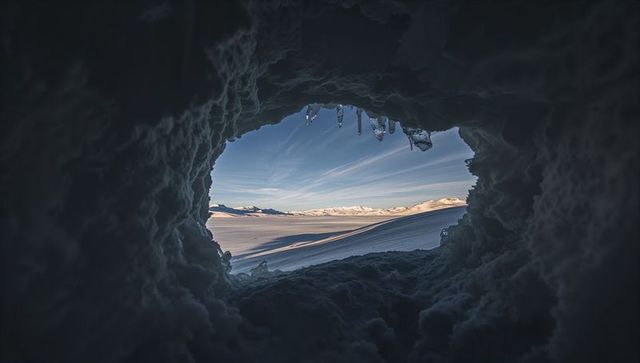 Snow cave framing sunlit alpine drifts and distant mountain horizon under icicle rim