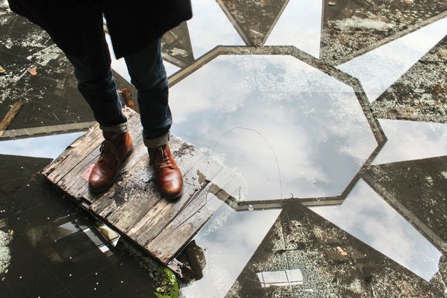Brown leather boots standing on wooden palate on patterned reflective floor. Person wearing jeans and coat gazing down at reflection. Perfect for use in urban exploration, fashion, boots advertisement or posters showing reflections and symmetry.