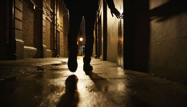 Shadowed pedestrian walking at night touching wet wall in urban alley, lamp reflections