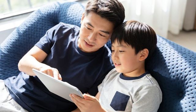 Father guiding son using tablet on blue sofa, sharing screen time, bonding and learning