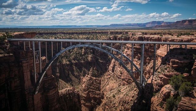 Spanning steel arch bridge rising over red sandstone canyon under dramatic sky