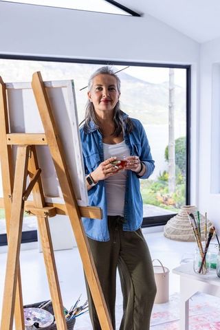 50s Woman Artist Standing Beside Easel Holding Mug Wearing Denim Shirt in Sunlit Studio