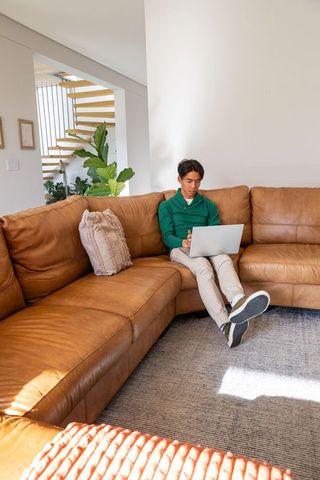 Asian man working on laptop on tan leather sectional sofa in sunlit modern living room