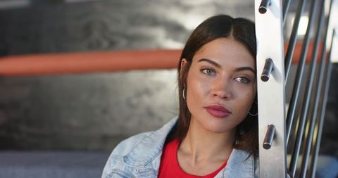 Indian woman leaning on metal railing in denim jacket and red top, moody lounge portrait