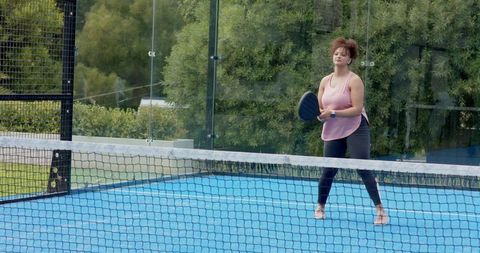 African american woman playing padel holding paddle on blue court with net and greenery
