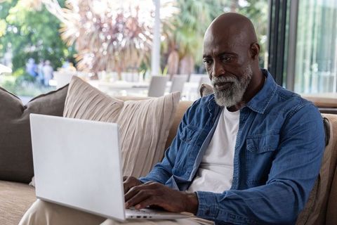 Mature african man working on laptop in sunlit living room near glass doors and patio