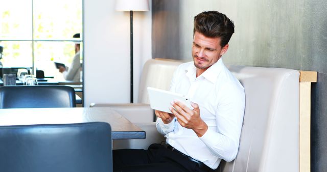 Businessman in white shirt using tablet in modern cafe. Ideal for topics on business technology, remote work, professional lifestyle, or modern work environments. Can be used in articles, blogs, marketing materials, or web design focusing on business, technology, and casual professional settings.