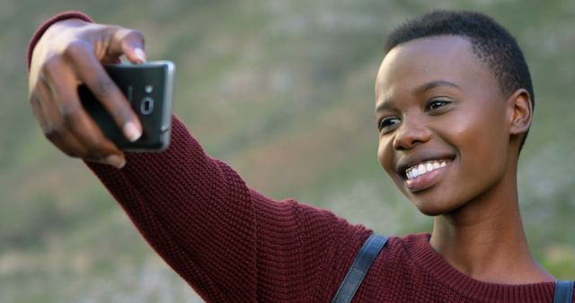 This image shows a happy young woman taking a selfie with her smartphone outdoors, capturing a joyful moment. She is wearing a casual sweater and has a backdrop of nature with blurred greenery. This can be used for advertisements promoting technology, lifestyle blogs, social media campaigns, or well-being articles.