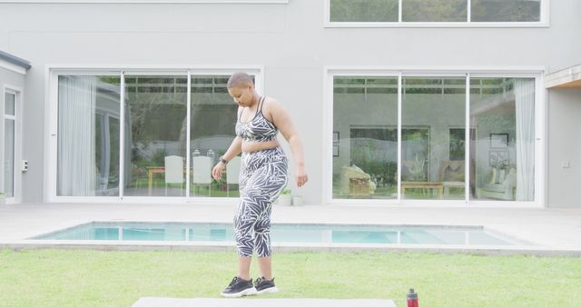 Adult woman engaged in a fitness routine near a modern home with a pool, demonstrating health and wellness lifestyle. Ideal for promoting fitness apparel, health and fitness blogs, outdoor exercise routines, or home workout products.