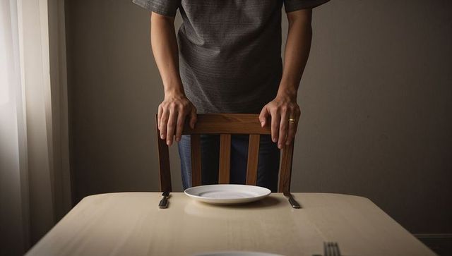 Standing man gripping chair back at dining table with empty plate, moody minimal interior