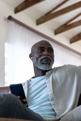 Senior african man relaxing on sofa holding smartphone wearing earbud in minimal home
