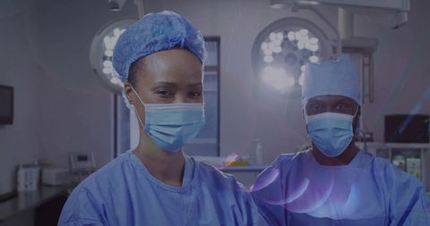 Two surgeons standing in operating room wearing masks and blue scrubs under surgical lights