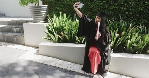 Indian woman graduate taking selfie in cap and gown holding diploma in sunlit garden