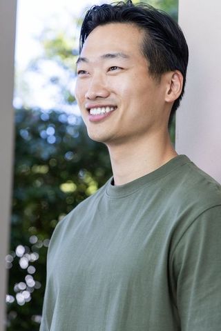 Asian man standing on porch smiling in olive tee with sunlit bokeh background