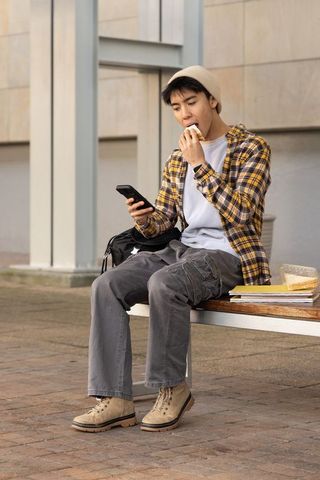 Asian male student eating sandwich while browsing smartphone on urban university bench