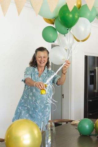 Senior woman decorating with green white gold balloons holding ribbons for home party