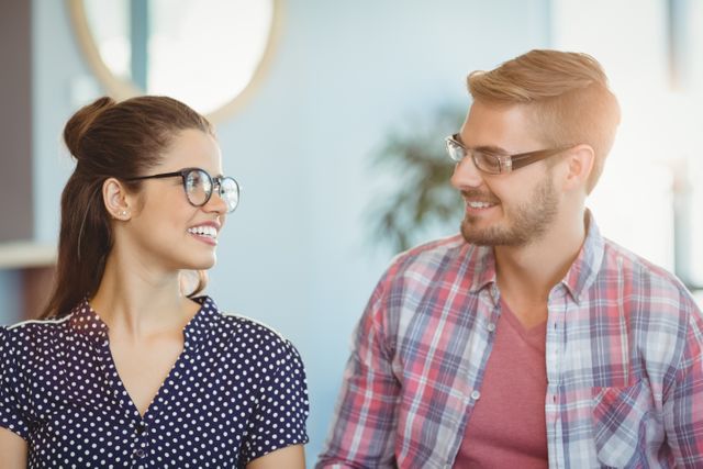 Young couple with glasses smiling and conversing in an office environment. Suitable for business and professional themes, coworking and teamwork concepts, or communication and collaboration visuals. Ideal for workplace culture promotions and corporate materials.