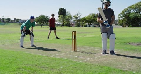 Batter gripping cricket bat at crease with wicketkeeper and fielder on grassy park pitch