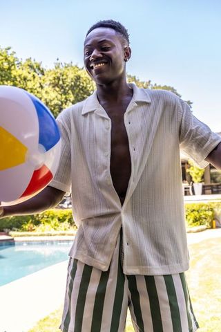 African american man smiling and tossing colorful beach ball by sunny backyard pool