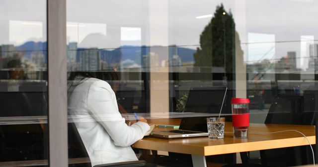 Businesswoman working at office table with a city skyline view reflected on the window. Image can be used for promoting urban workspaces, business productivity, corporate lifestyle, modern office designs, professional work settings.
