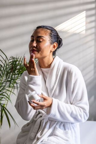 Asian woman applying moisturizer in sunlit white robe for calm morning skincare routine