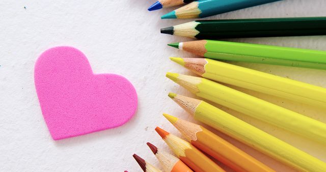 A pink heart-shaped sponge lies next to a gradient of green to yellow colored pencils on a white background, with copy space. The arrangement suggests creativity and could be associated with art, education, or crafting activities.