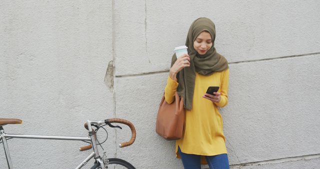 Woman in hijab standing against wall holding paper coffee cup and smartphone while smiling. Bicycle parked beside her. Perfect for depicting diversity, urban lifestyles, and technology use.