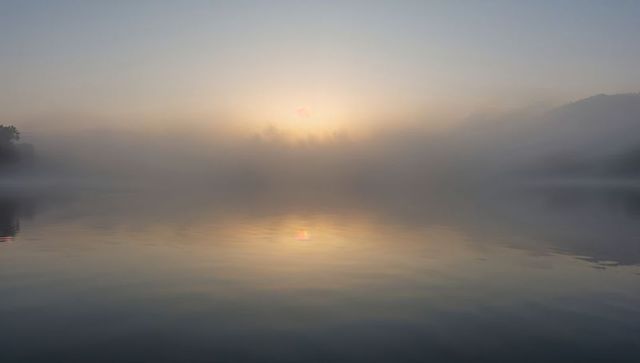Glowing mist covering lake reflecting pale sunrise over calm mirrorlike water and treeline