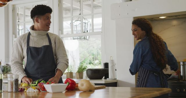 Couple is preparing food in their bright, modern kitchen. Both are wearing aprons and seem engaged in the cooking process with fresh vegetables displayed on the counter. This scene can be used for themes related to relationships, home life, cooking together, healthy living, or culinary activities.
