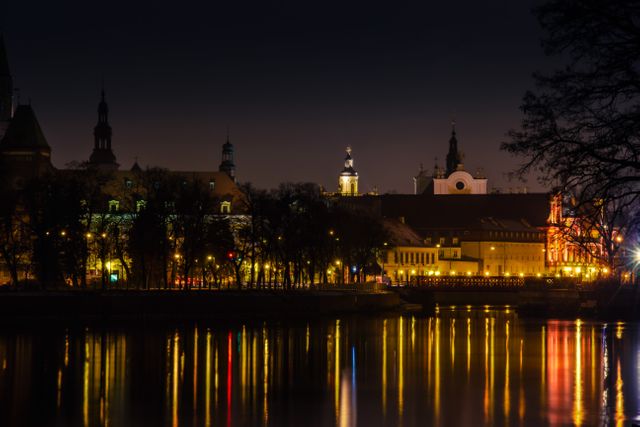 Historic city skyline reflecting in calm river during night. Golden lights and silhouettes of church spires and architecture create a serene scene. Suitable for travel guides, wallpapers, and articles promoting urban tourism and night photography.