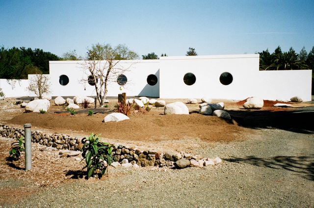 Modern white building features circular windows, minimalist architecture style with desert-inspired landscaping. Ideal use for contents focused on contemporary design, architecture, landscaping insights, beach house inspirations, and exterior decoration.