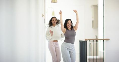 Diverse female friends dancing down bright home hallway in loungewear celebrating morning