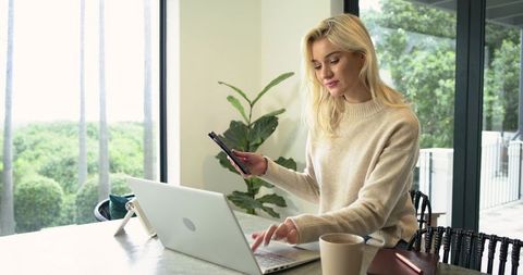 Blonde woman working remotely from bright home office with laptop smartphone and coffee
