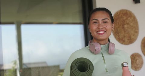 Smiling asian woman holding yoga mat and water bottle wearing headphones and mint activewear