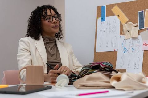 African american fashion designer arranging fabric samples at table viewing corkboard sketches