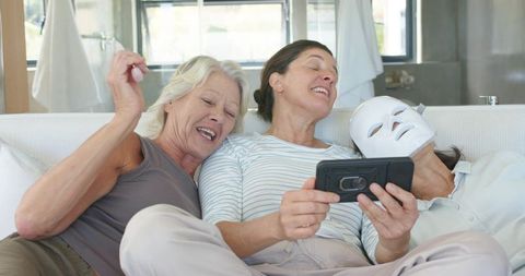 Senior women laughing, sharing smartphone, using led facial mask for skincare night