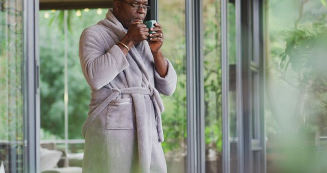 Mature man standing near large windows, drinking coffee, wearing bathrobe, relaxing at home. Image can be used for concepts of morning routine, home comfort, leisure activities, peaceful environment, and lifestyle.