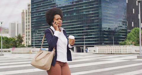 Professional woman walking and talking on phone holding coffee and handbag in modern plaza