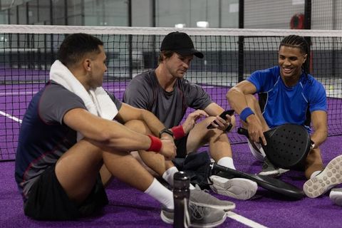 Three male padel players sitting on purple court checking smartphone after match break