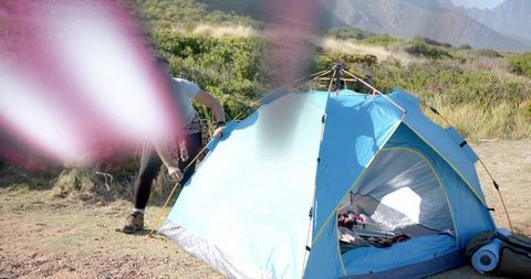 Mid-adult woman pitching blue dome tent on coastal grassland, backpack and camping gear