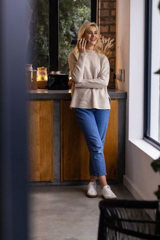 Blonde woman chatting on smartphone while leaning at cozy modern kitchen counter by window and plant