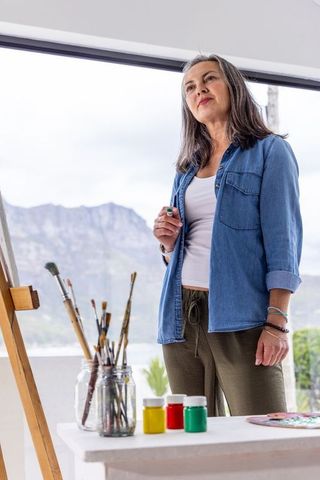 Mature artist standing by easel in bright studio overlooking mountain lake, holding brush