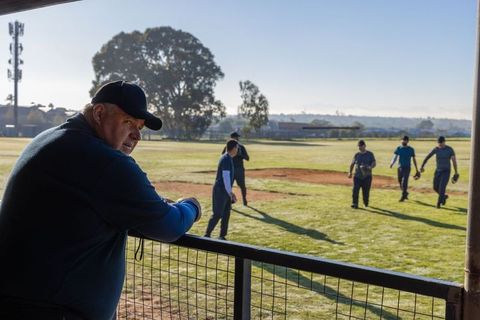 Senior man watching amateur baseball practice from dugout fence in morning sunlight