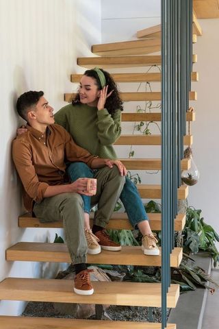Young couple sitting on floating wooden stairs holding ceramic mug among indoor plants