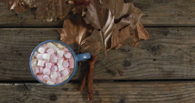 Hot chocolate with marshmallows and cinnamon sticks surrounded by dried autumn leaves on a rustic wooden table creates a cozy and seasonal atmosphere. Perfect for advertising fall recipes, comfort food, or autumn celebrations. Suitable for use in blogs, social media, or seasonal advertising campaigns.