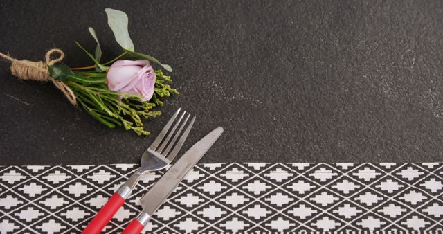 A single pink rose tied with twine lies next to a fork and knife on a patterned tablecloth, with copy space. This setting suggests a romantic meal or a special occasion dining experience.